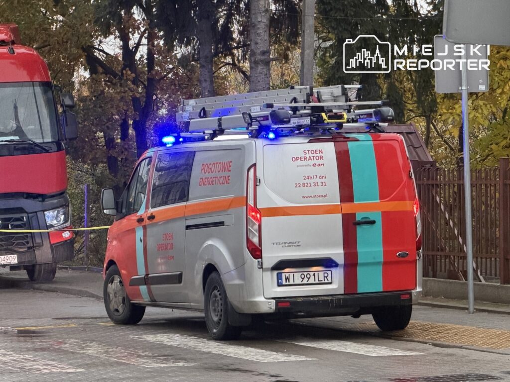 Samochód pogotowia energetycznego z włączonymi niebieskimi światłami stoi na ulicy obok czerwonego pojazdu ciężarowego.