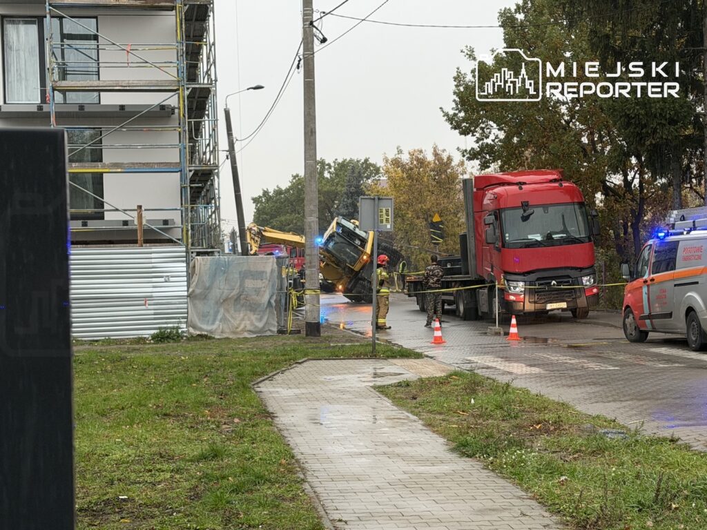 Przewrócona koparka przy ulicy, obok ciężarówka i służby ratunkowe zabezpieczające miejsce zdarzenia.