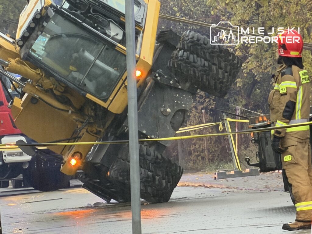 Przewrócona żółta koparka na ulicy, obok strażak i taśma zabezpieczająca miejsce zdarzenia.