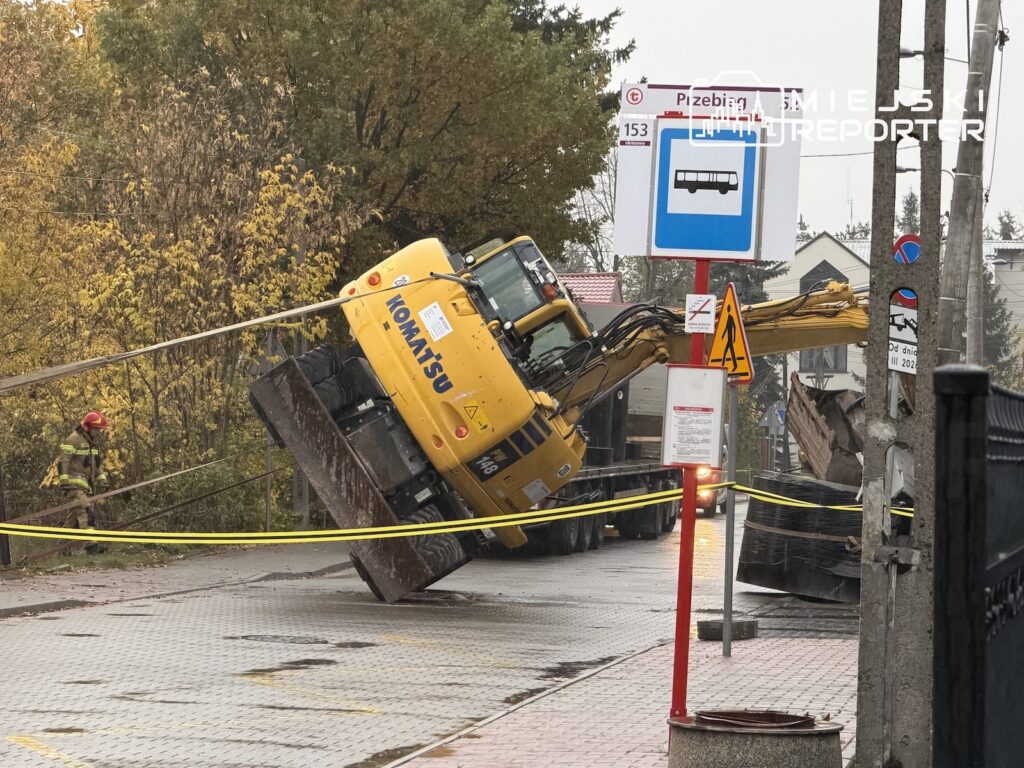 Żółta koparka przewrócona na bok przy przystanku autobusowym, otoczona taśmą zabezpieczającą.