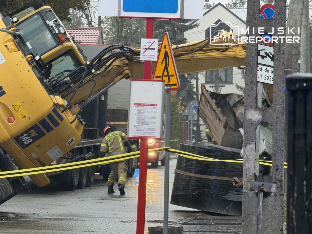 Przewrócona żółta koparka leży obok słupa, a w pobliżu pracuje ratownik; miejsce zabezpiecza żółta taśma.
