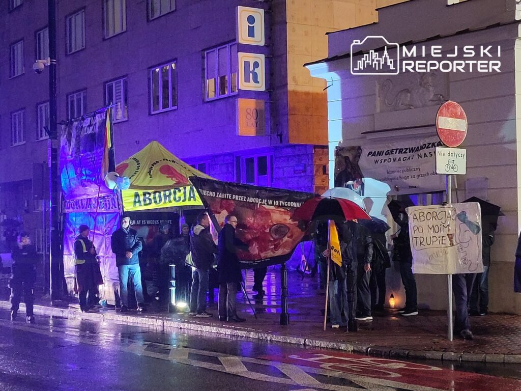 Protestujący stoją nocą na mokrym chodniku pod banerem z napisem „ABORCJA”, w świetle niebieskich policyjnych świateł.