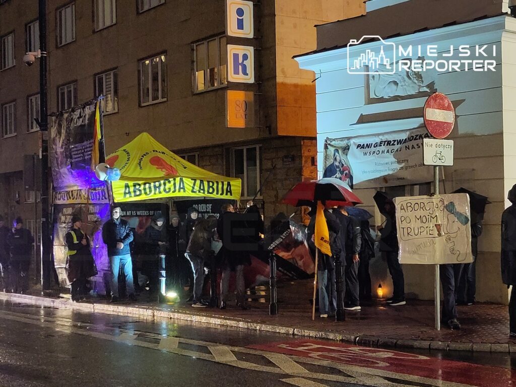 Grupa osób z parasolami protestuje nocą pod żółtym namiotem z napisem „ABORCJA ZABIJA”, przy mokrej ulicy.