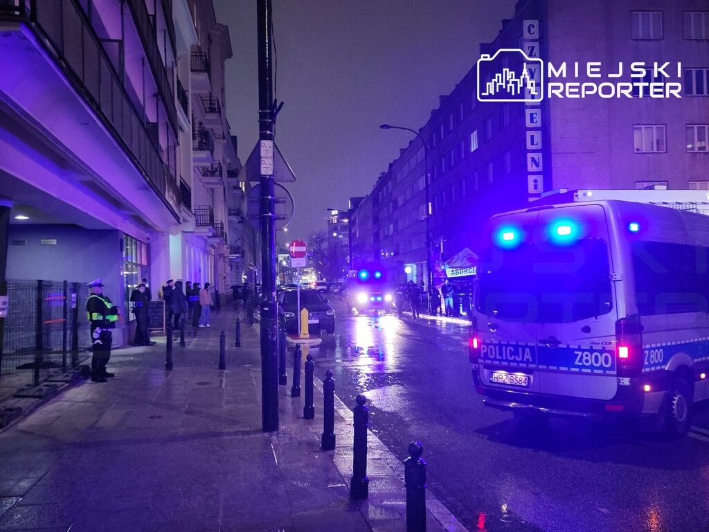 Policyjny bus z włączonymi niebieskimi światłami stoi na mokrej ulicy, w tle funkcjonariusze i piesi nocą.