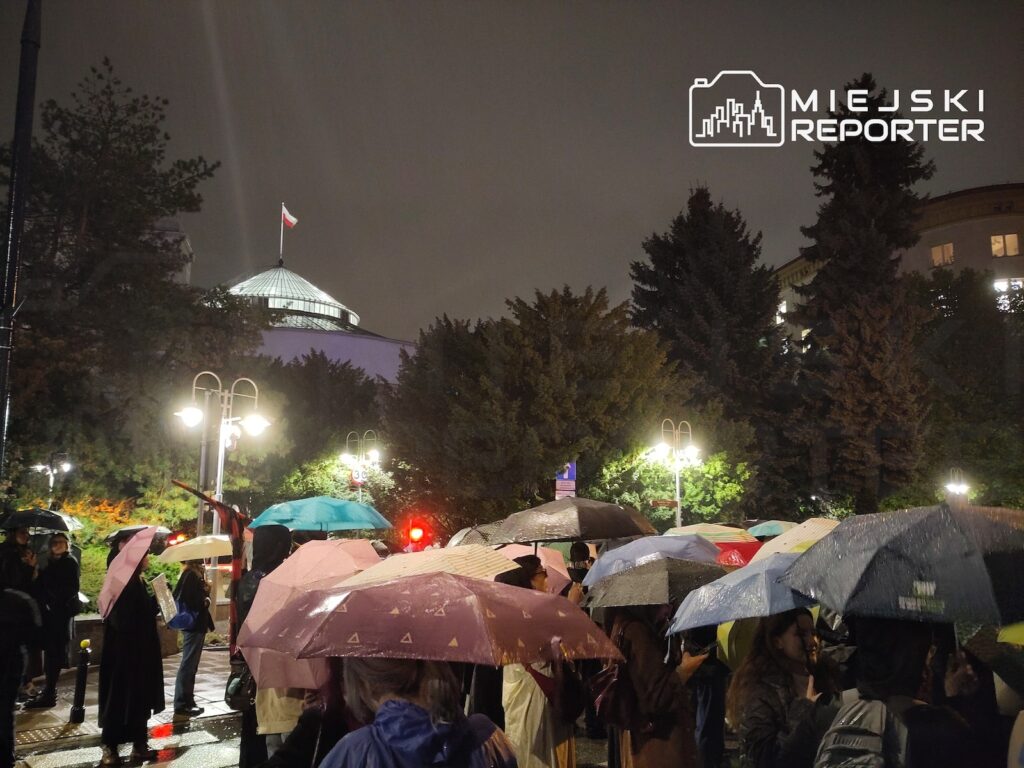 Protestujący stoją pod parasolami nocą na mokrym placu, w tle oświetlona kopuła budynku z polską flagą.