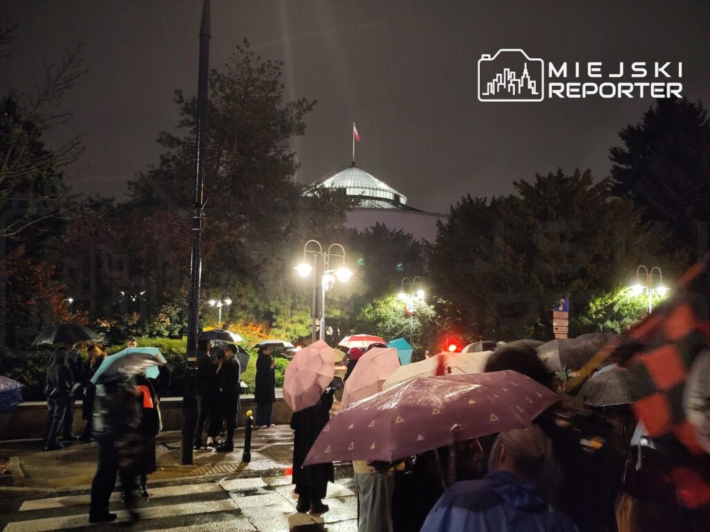 Tłum ludzi z parasolami stoi w deszczu na oświetlonym placu przed budynkiem z kopułą i polską flagą.