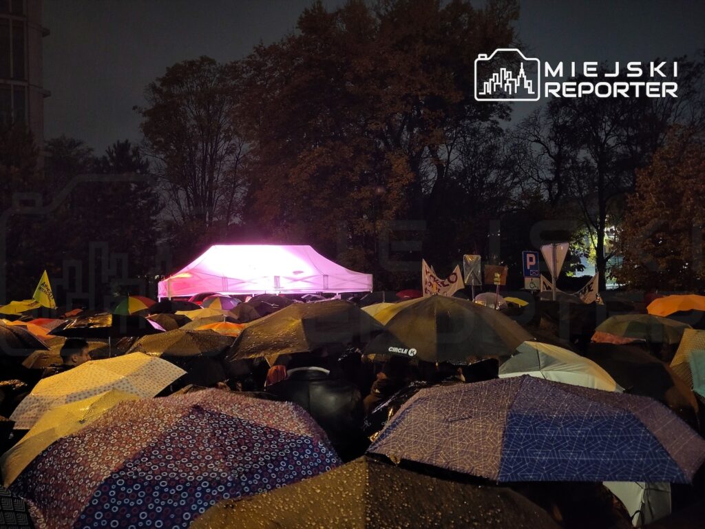 Tłum ludzi zebranych pod kolorowymi parasolami wokół podświetlonego namiotu po zmroku, wśród drzew