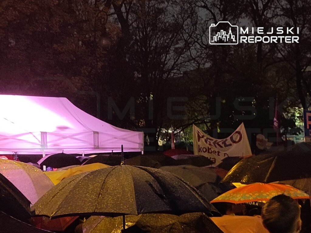 Protestujący stoją pod parasolami w deszczu obok oświetlonego namiotu i transparentu z napisem „Piekło kobiet”.