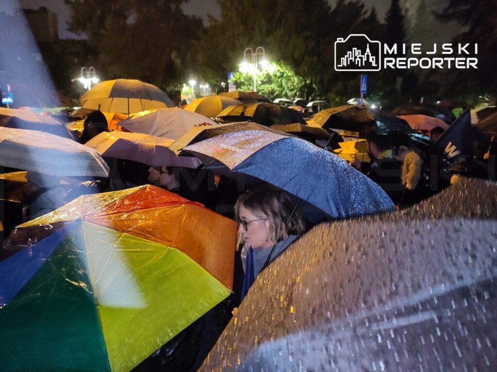 Tłum ludzi stoi nocą pod kolorowymi parasolami w deszczu, wśród oświetlonych drzew.