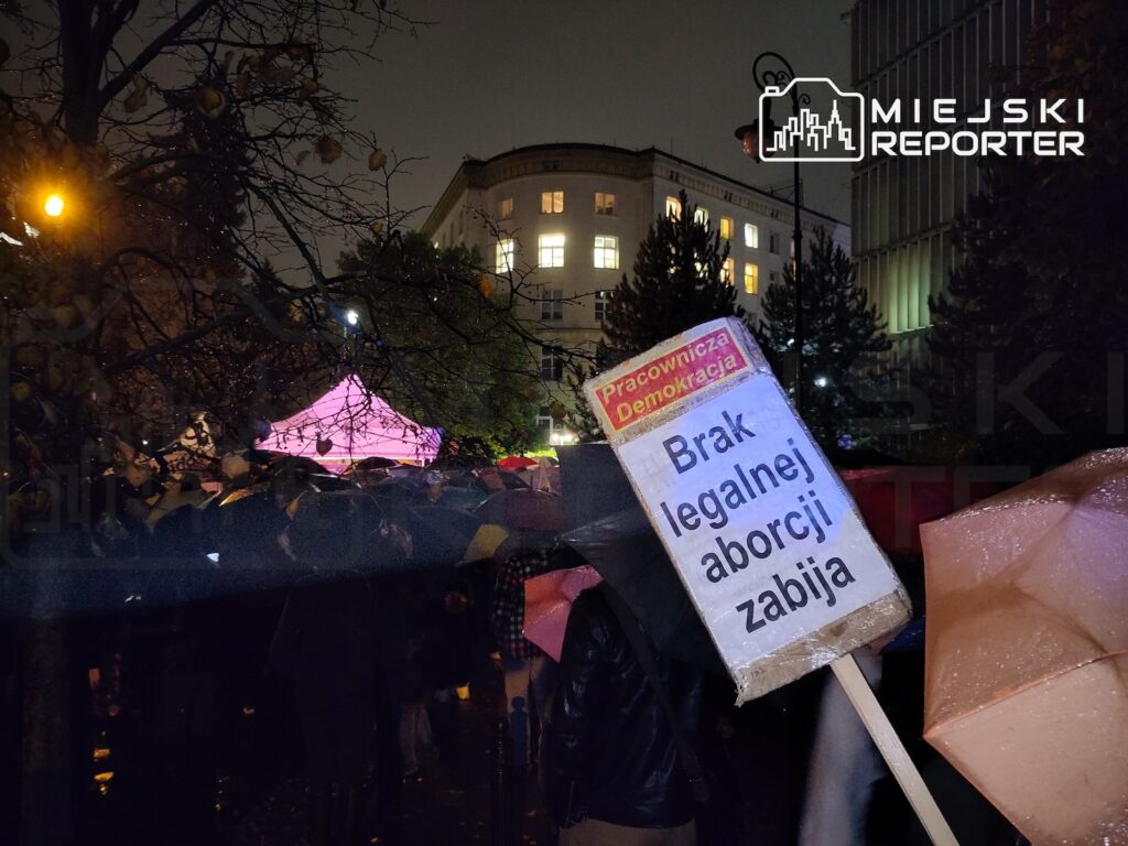 Protestujący trzymający transparent „Brak legalnej aborcji zabija” w tłumie pod parasolami, nocą przy oświetlonych budynkach.