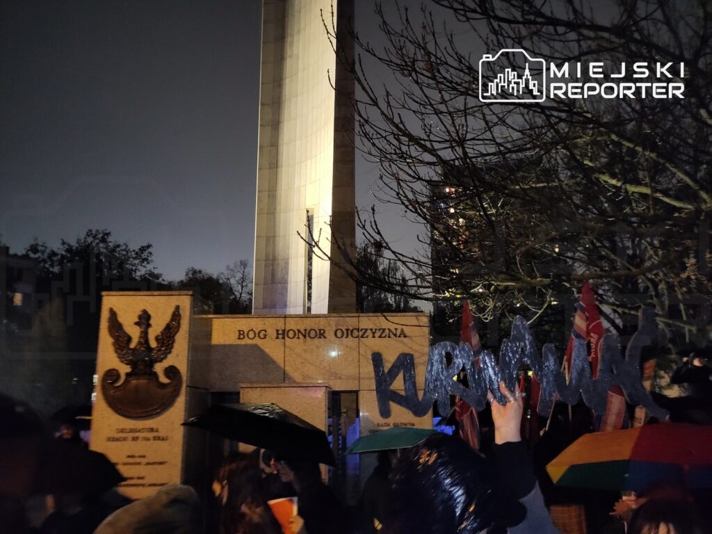 Tłum protestujących z transparentami i flagami stoi przy murze z napisem „BÓG HONOR OJCZYZNA”, nocą podświetlonym od góry.