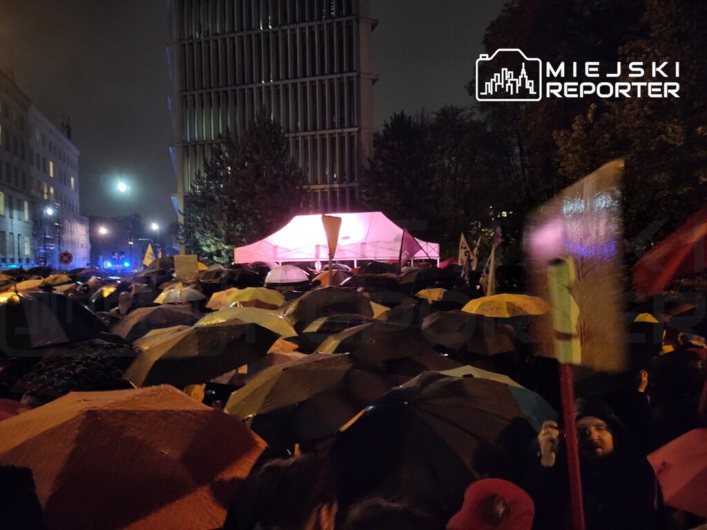 Tłum ludzi z parasolami zgromadzony nocą wokół podświetlonego namiotu między miejskimi budynkami.