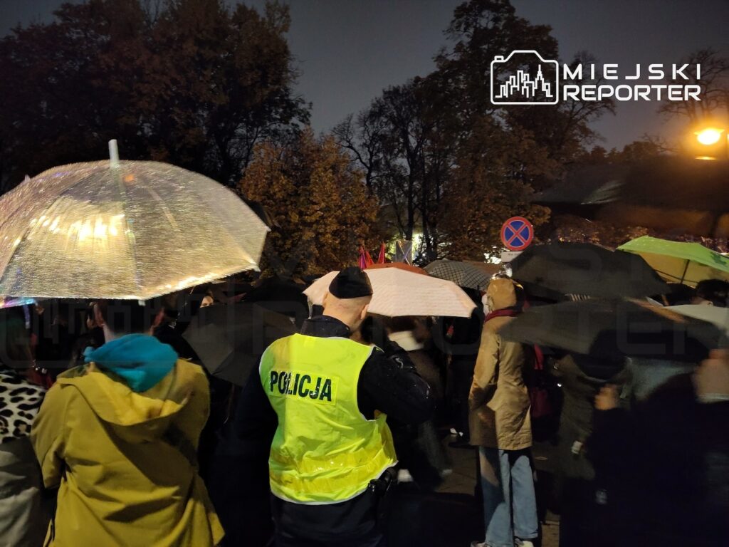 Policjant w odblaskowej kamizelce stoi wśród osób z parasolami na ciemnej, deszczowej ulicy.