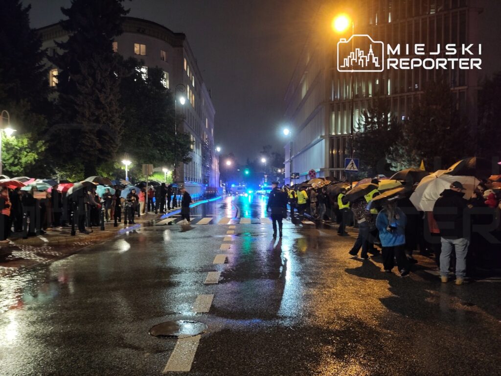 Dwie grupy ludzi z parasolami stoją po obu stronach mokrej ulicy, a w oddali świecą niebieskie światła pojazdów policyjnych.
