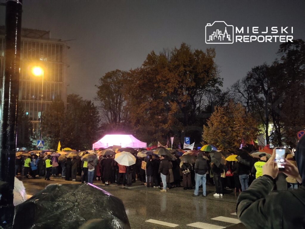 Tłum ludzi z parasolami gromadzi się nocą w deszczu przy oświetlonym namiocie, a osoba na pierwszym planie robi zdjęcie