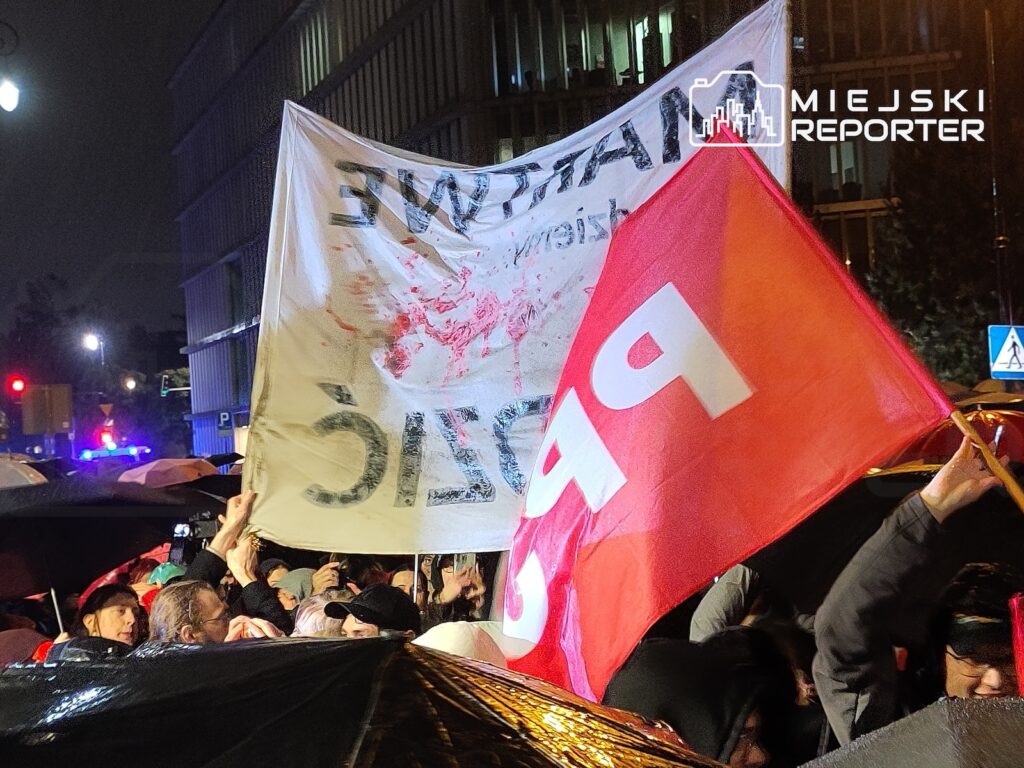 Tłum protestujących trzyma biały transparent z odwróconym napisem i czerwono-białą flagę przed oświetlonym nocą budynkiem.