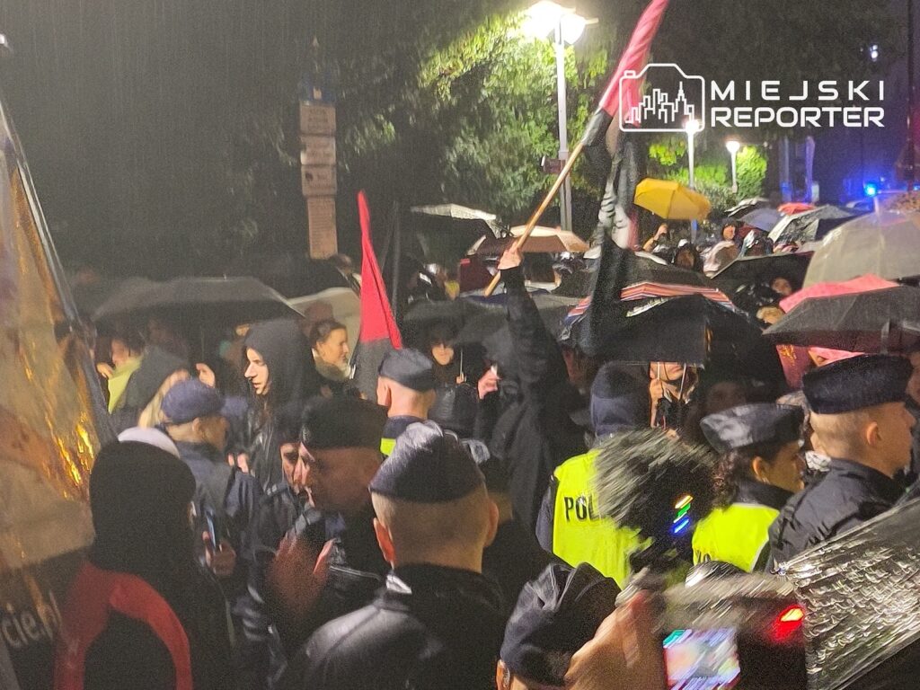 Policjanci w kamizelkach odblaskowych stoją w tłumie ludzi z parasolami i flagami, nocą na ulicy.