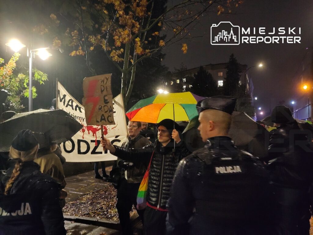 Policjanci stoją przed grupą protestujących z transparentami pod kolorowymi parasolami podczas nocnej demonstracji w deszczu.