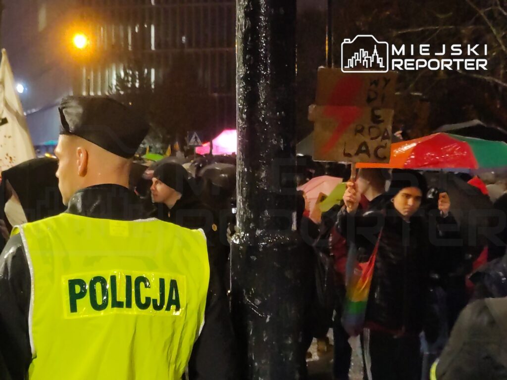 Policjant w odblaskowej kamizelce stoi przed grupą protestujących z parasolami i kartonowym transparentem nocą.