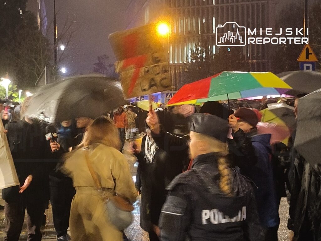 Uczestnicy protestu z parasolami i transparentem stoją nocą na mokrej ulicy, obok policjantki tyłem do aparatu.