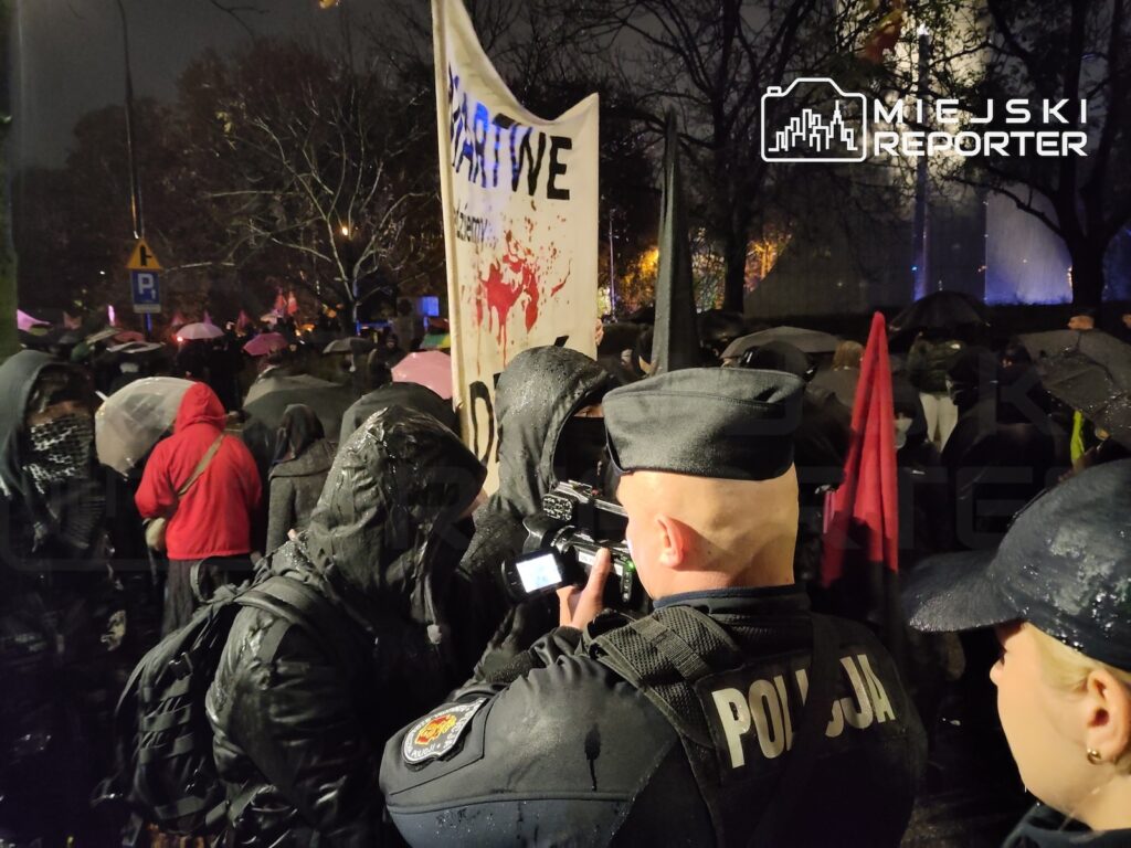 Policjanci w czarnych mundurach stoją w tłumie osób z parasolami obok protestowego transparentu nocą.