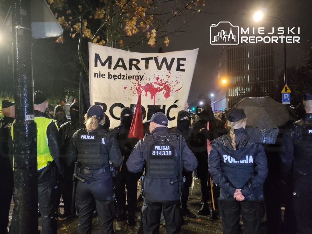 Policjanci w mundurach stoją przed protestującymi z transparentem „MARTWE nie będziemy” na ulicy nocą.