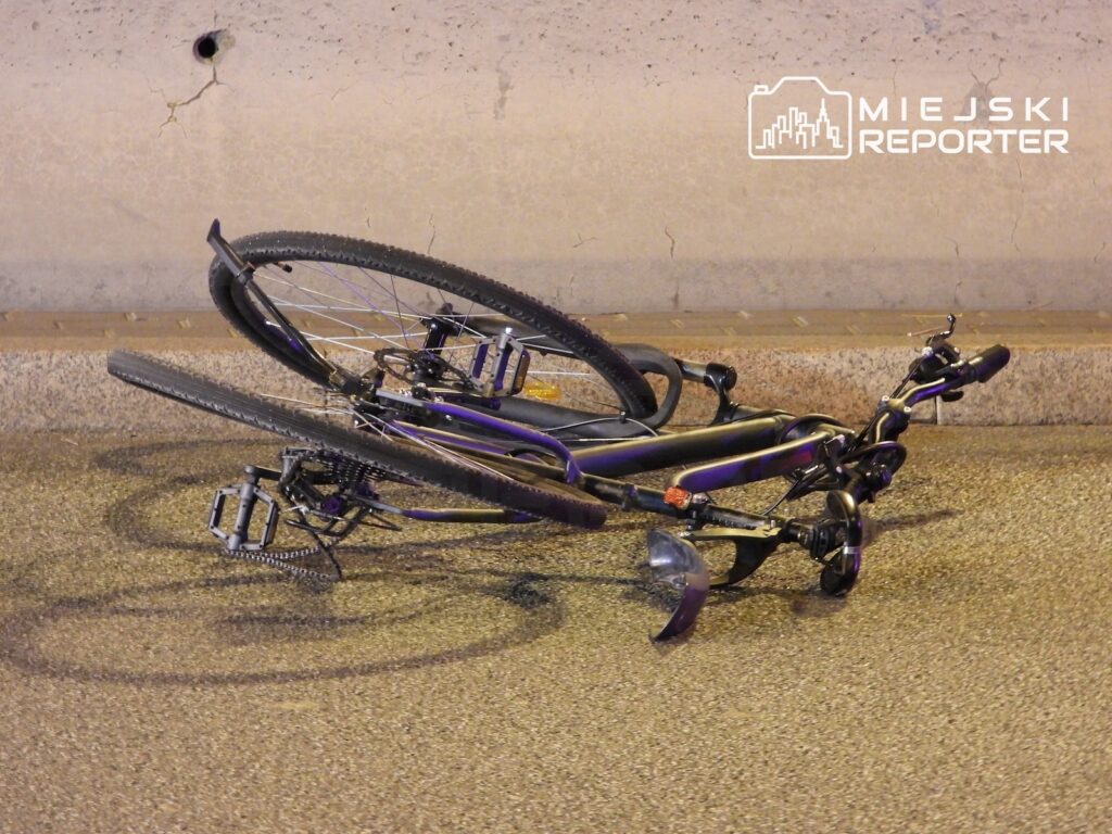 Przewrócony czarno-fioletowy rower leży na jezdni przy betonowej ścianie.