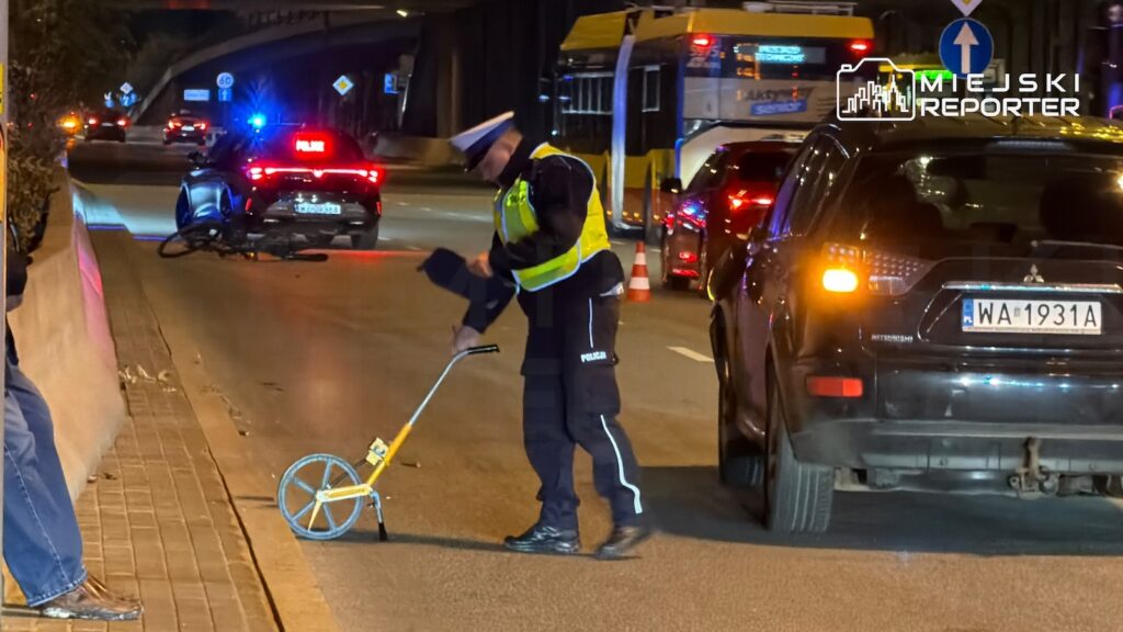 Policjant w kamizelce odblaskowej mierzy odległość na jezdni przy radiowozie i autobusie nocą.
