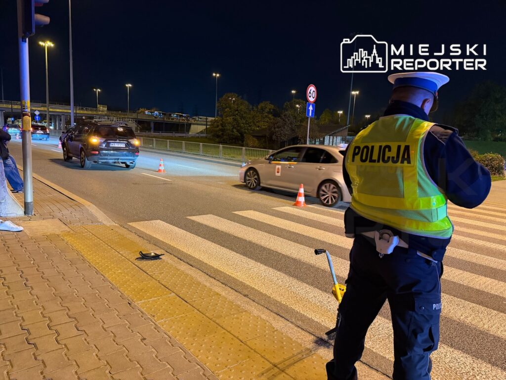 Policjant w kamizelce odblaskowej kieruje ruchem przy przejściu dla pieszych, obok stoją samochody i pachołki nocą.