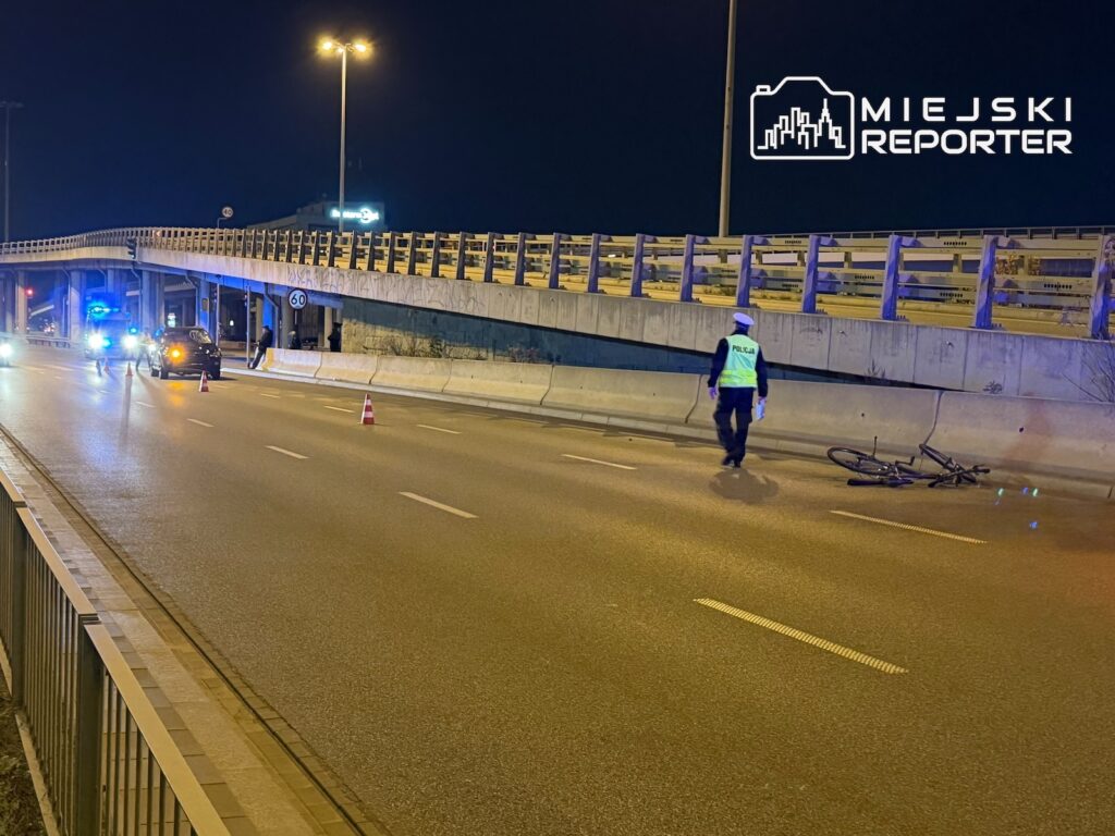 Policjant w odblaskowej kamizelce stoi obok przewróconego roweru na nocnej ulicy, przy zablokowanym pasie ruchu.