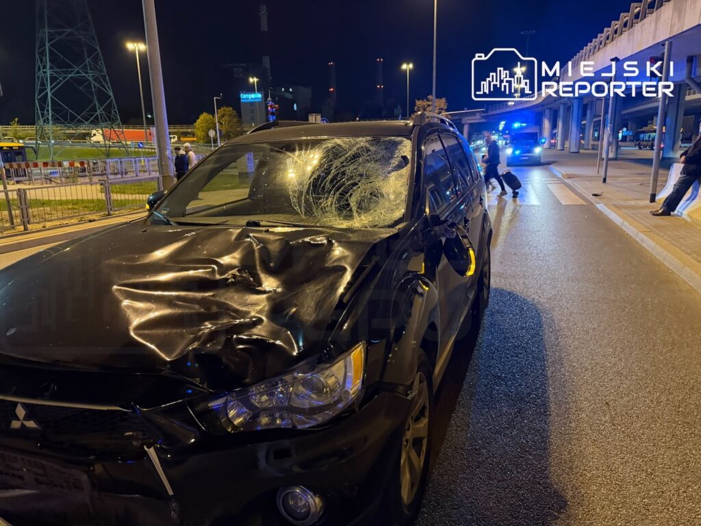 Rozbity czarny SUV z wgniecioną maską i pękniętą szybą stoi nocą na ulicy, w tle świecą światła służb.