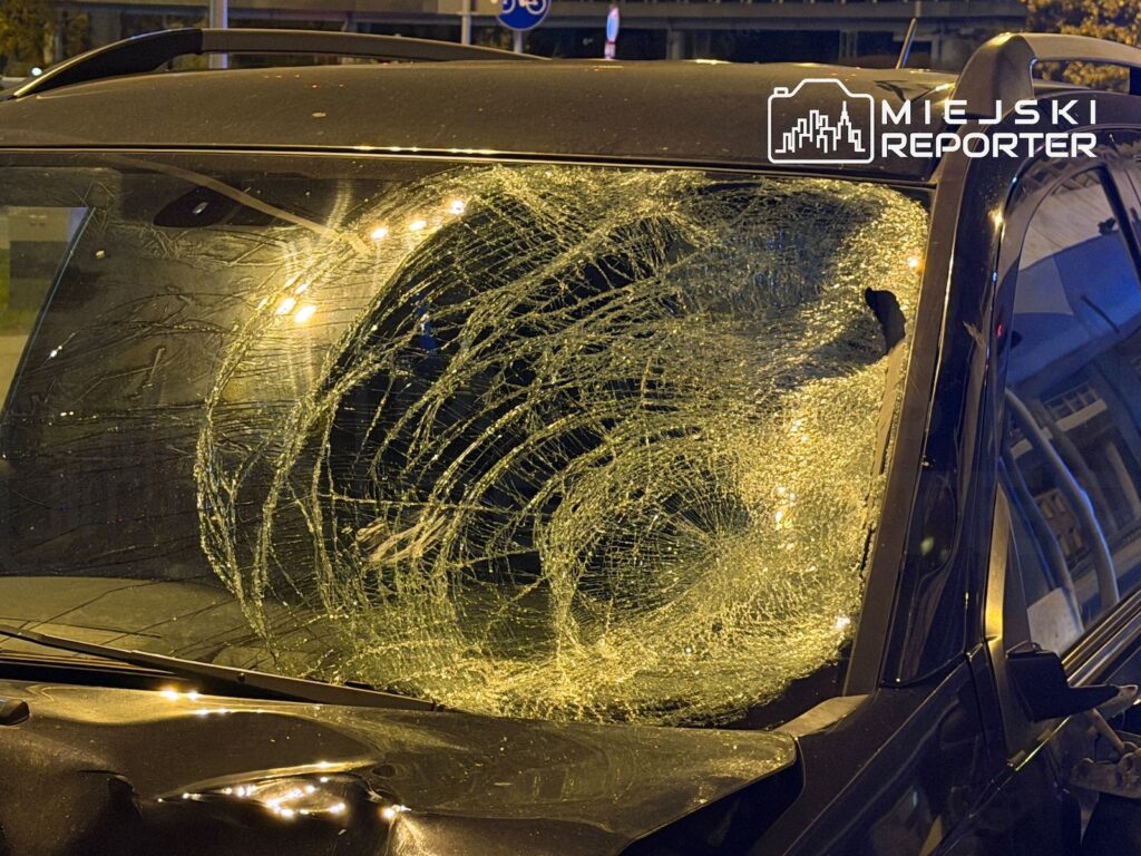 Samochód z mocno rozbitą przednią szybą stoi nocą na oświetlonej ulicy.