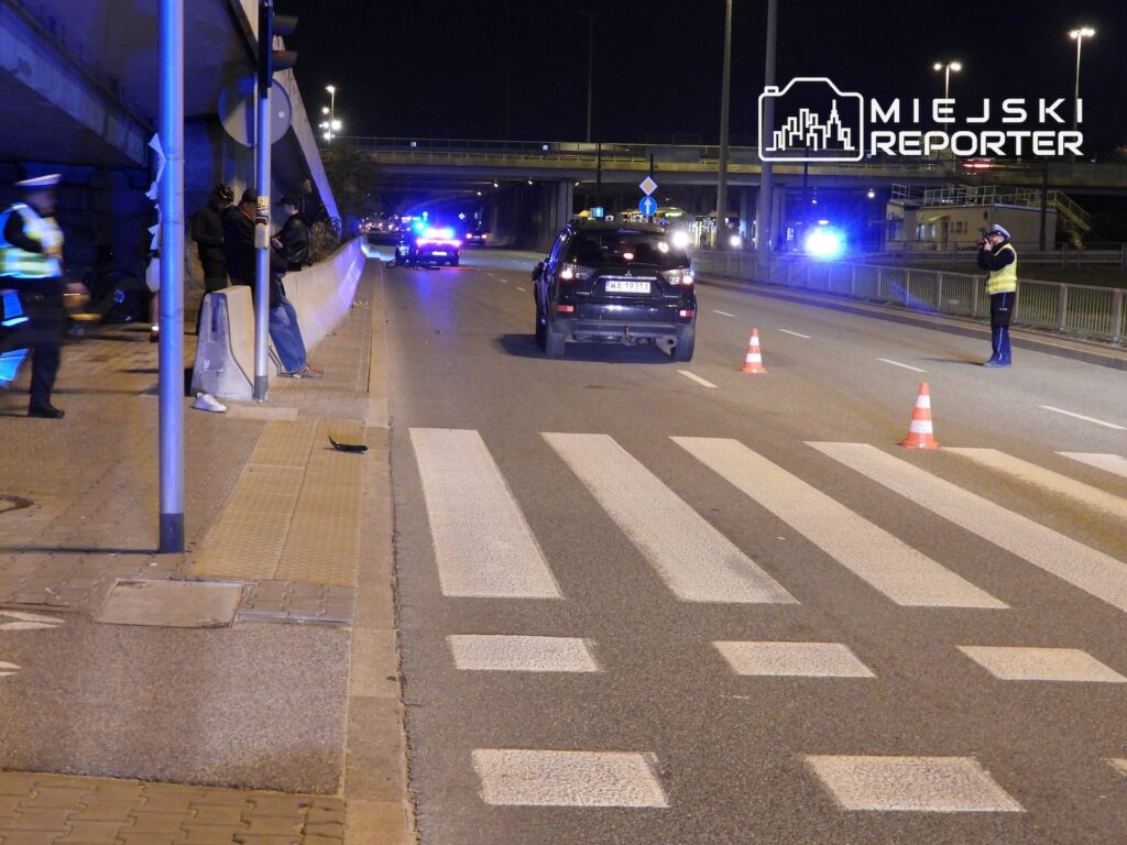 Radiowóz i samochód stoją na nocnej jezdni przy przejściu dla pieszych, zabezpieczonej pachołkami i przez policjanta.