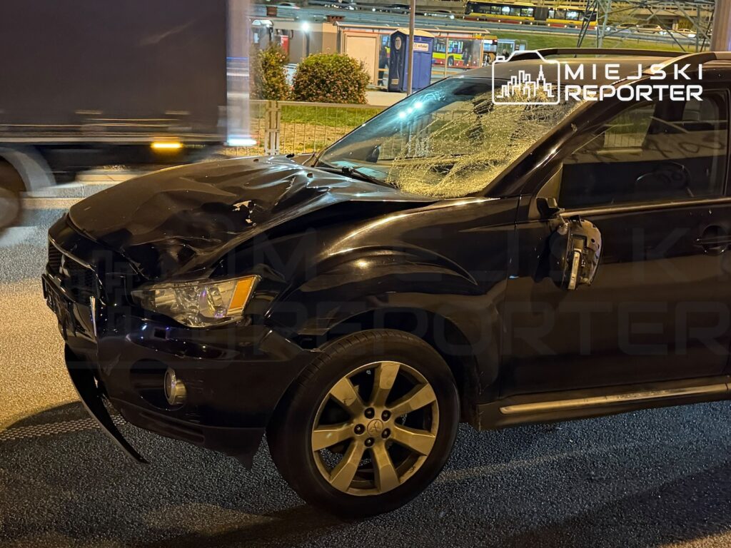 Uszkodzony czarny samochód z wgniecioną maską i rozbitą przednią szybą stoi nocą przy ulicy.