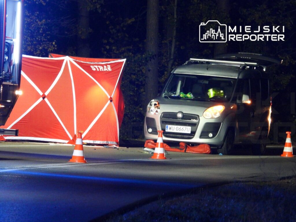 Srebrny samochód dostawczy stoi na nocnej drodze, otoczony pachołkami i czerwonym parawanem strażackim.