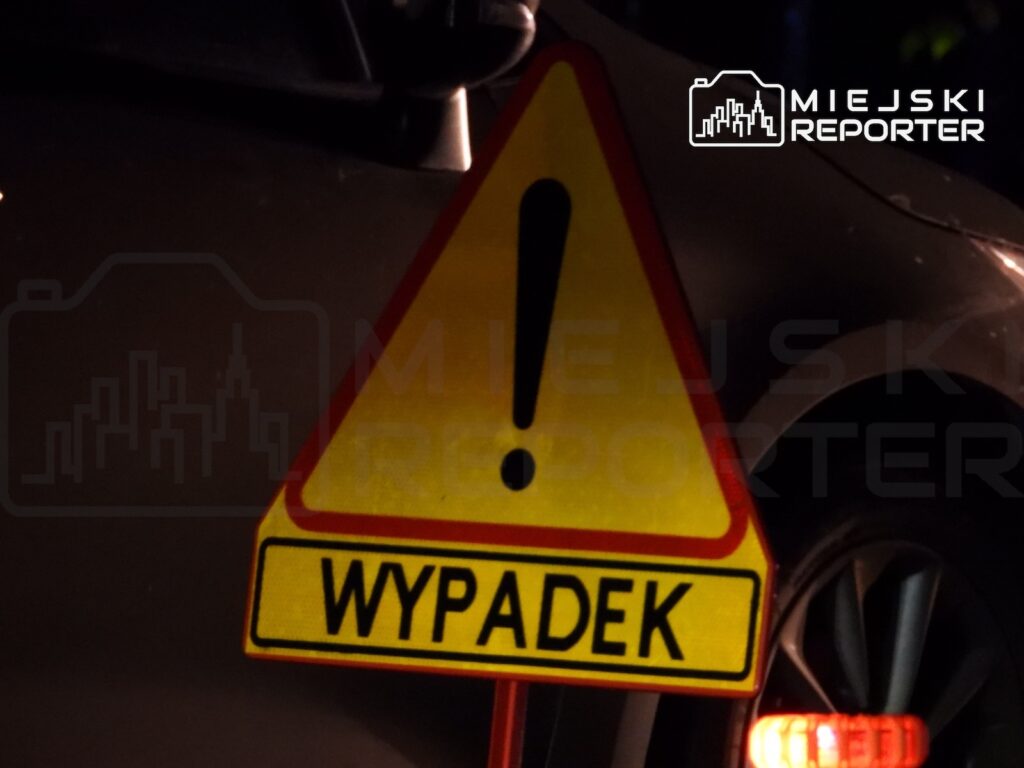 Żółty znak ostrzegawczy z wykrzyknikiem i napisem „WYPADEK” ustawiony przy samochodzie nocą.