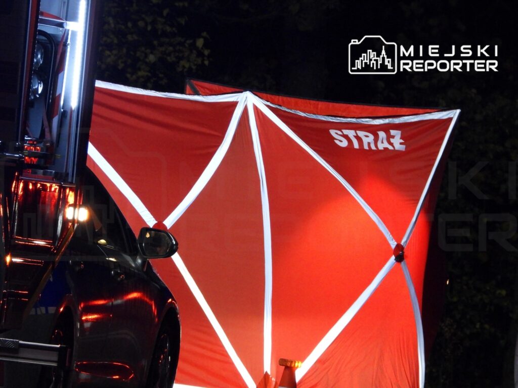 Czerwony parawan z napisem „STRAŻ” ustawiony obok pojazdu strażackiego nocą.