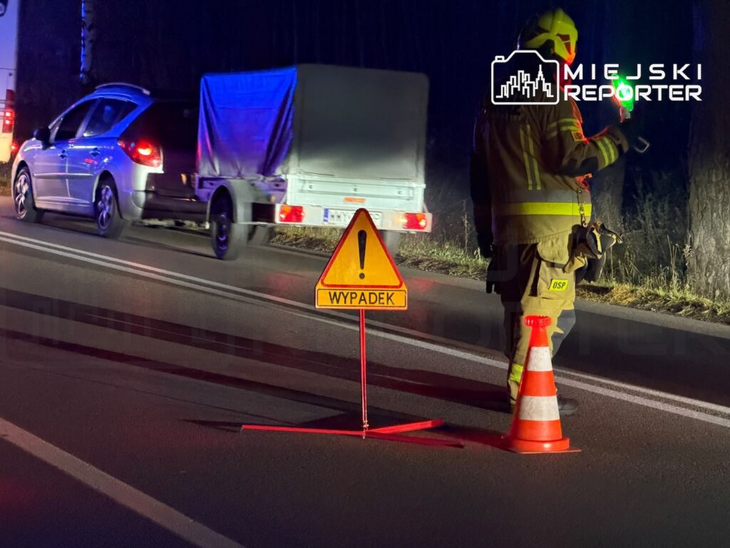Strażak w odblaskowym ubraniu zabezpiecza nocą miejsce wypadku na drodze; obok stoi znak „WYPADEK” i pachołek.