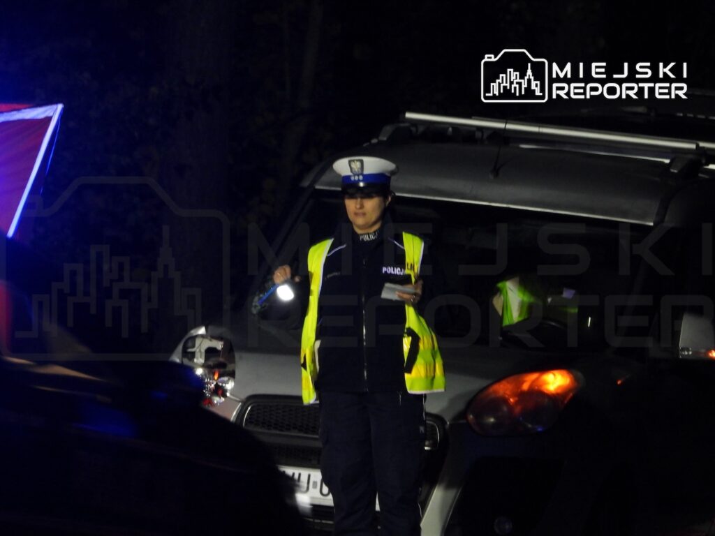 Policjantka w odblaskowej kamizelce kieruje ruchem nocą przed radiowozem i innymi samochodami.
