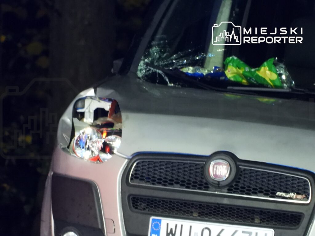 Przód samochodu Fiat z rozbitym lewym reflektorem i uszkodzoną przednią szybą, zaparkowanego po zmroku.
