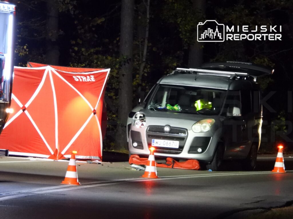 Samochód osobowy stoi nocą na jezdni przy rozstawionym czerwonym parawanie straży pożarnej i pachołkach drogowych.