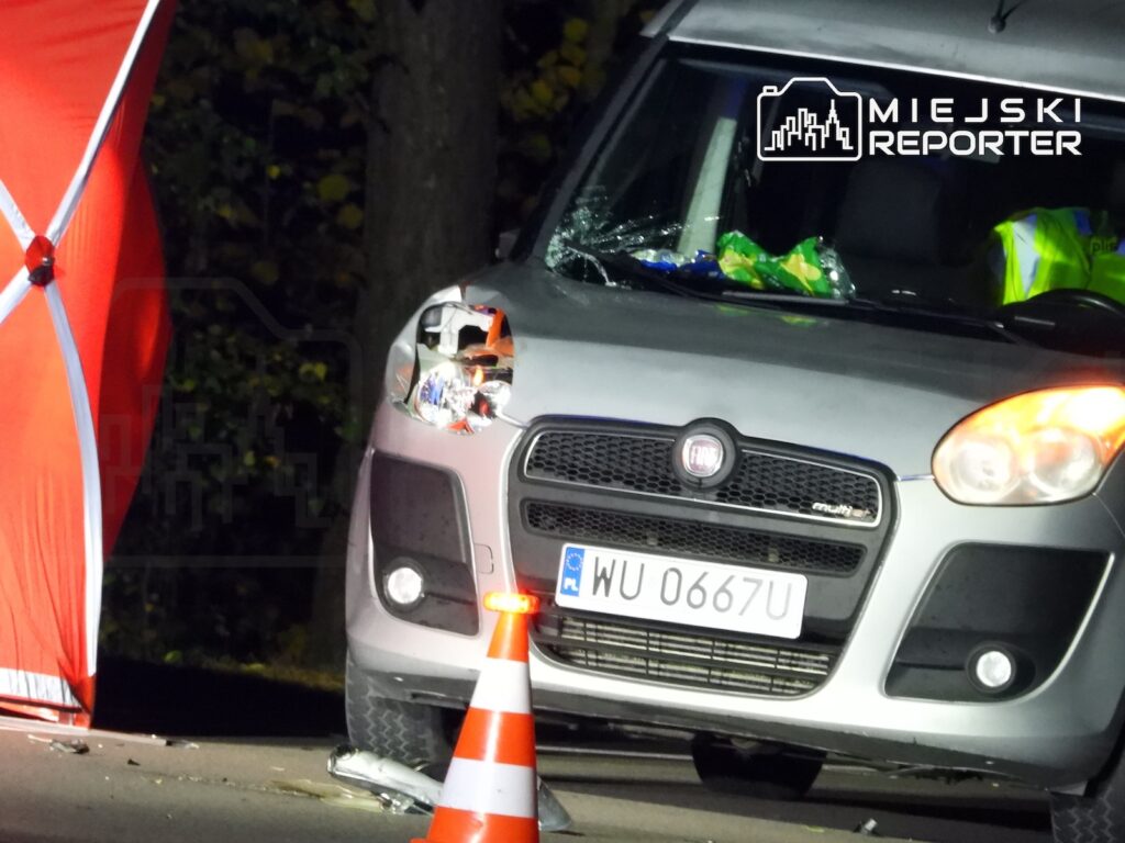 Uszkodzony przód srebrnego Fiata z rozbitym lewym reflektorem, zabezpieczony pachołkiem obok czerwonego namiotu