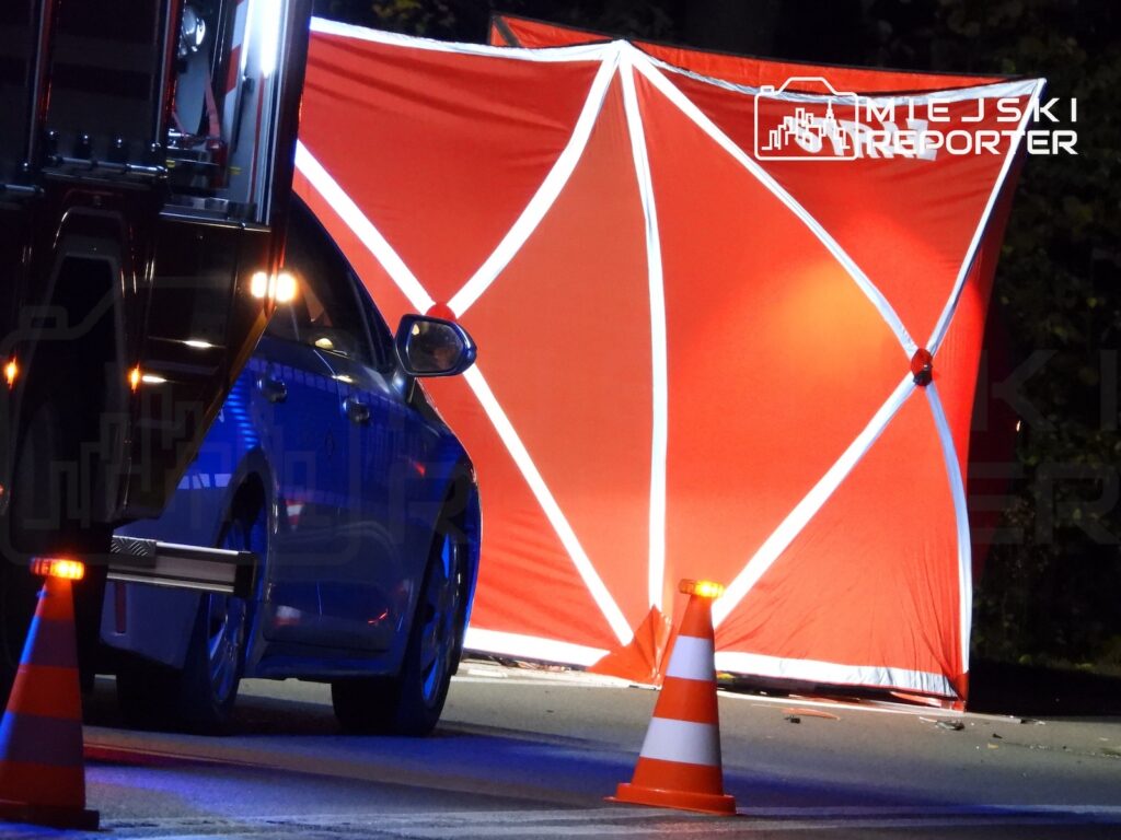 Pomarańczowy parawan odgradza miejsce zdarzenia na nocnej ulicy, obok stoją radiowóz i pachołki drogowe.