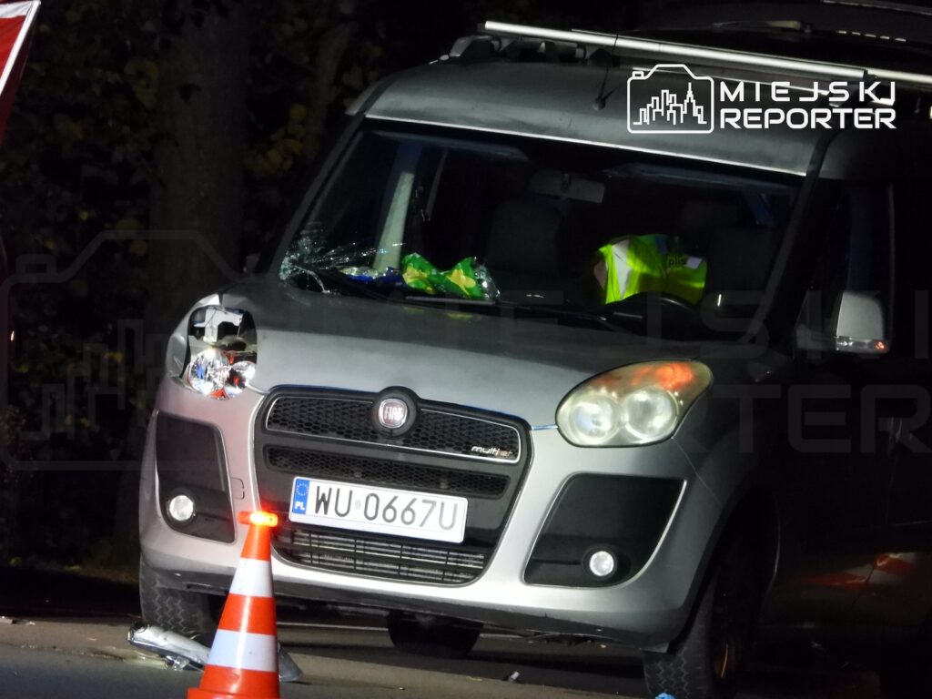 Uszkodzony srebrny Fiat stoi nocą na drodze, przed nim znajduje się pomarańczowo-biały pachołek drogowy.