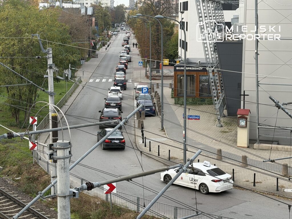Długi sznur samochodów stoi na ulicy przy torowisku tramwajowym, obok nowoczesnego budynku biurowego.