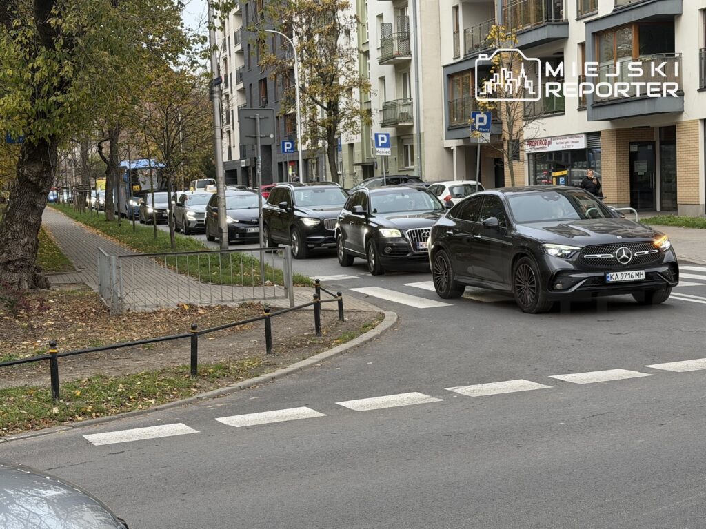 Kolejka samochodów stoi na ulicy przy przejściu dla pieszych, między blokami mieszkalnymi a chodnikiem z drzewami.