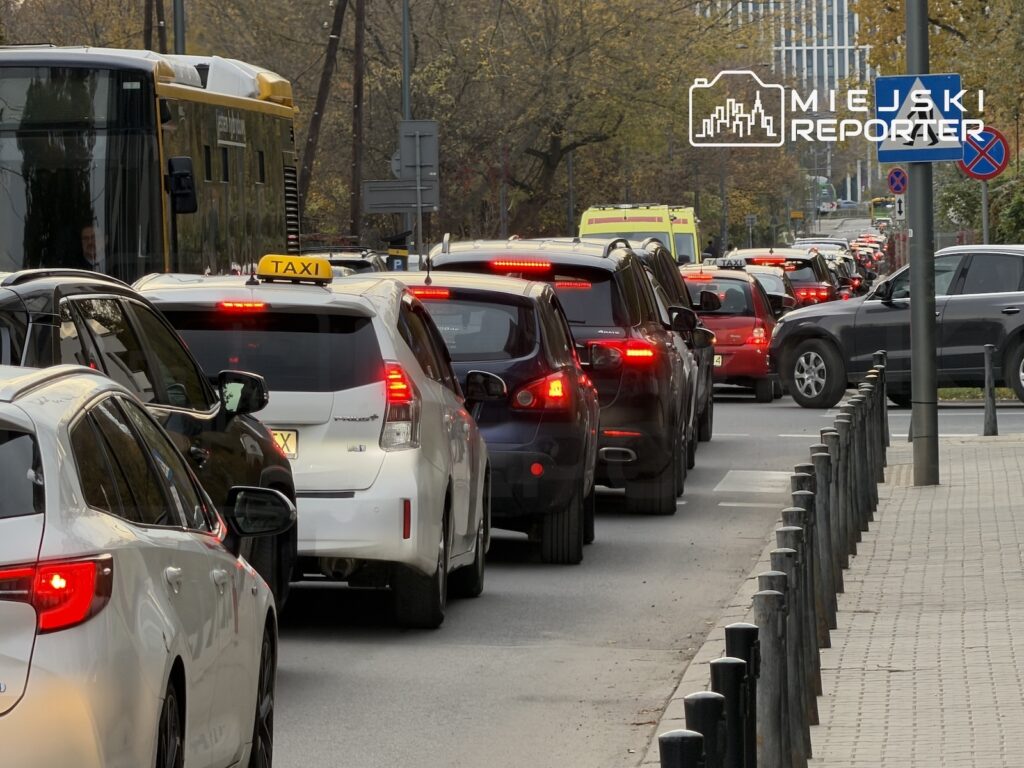 Długi sznur samochodów z zapalonymi światłami stopu stoi w korku obok autobusu miejskiego i karetki pogotowia.