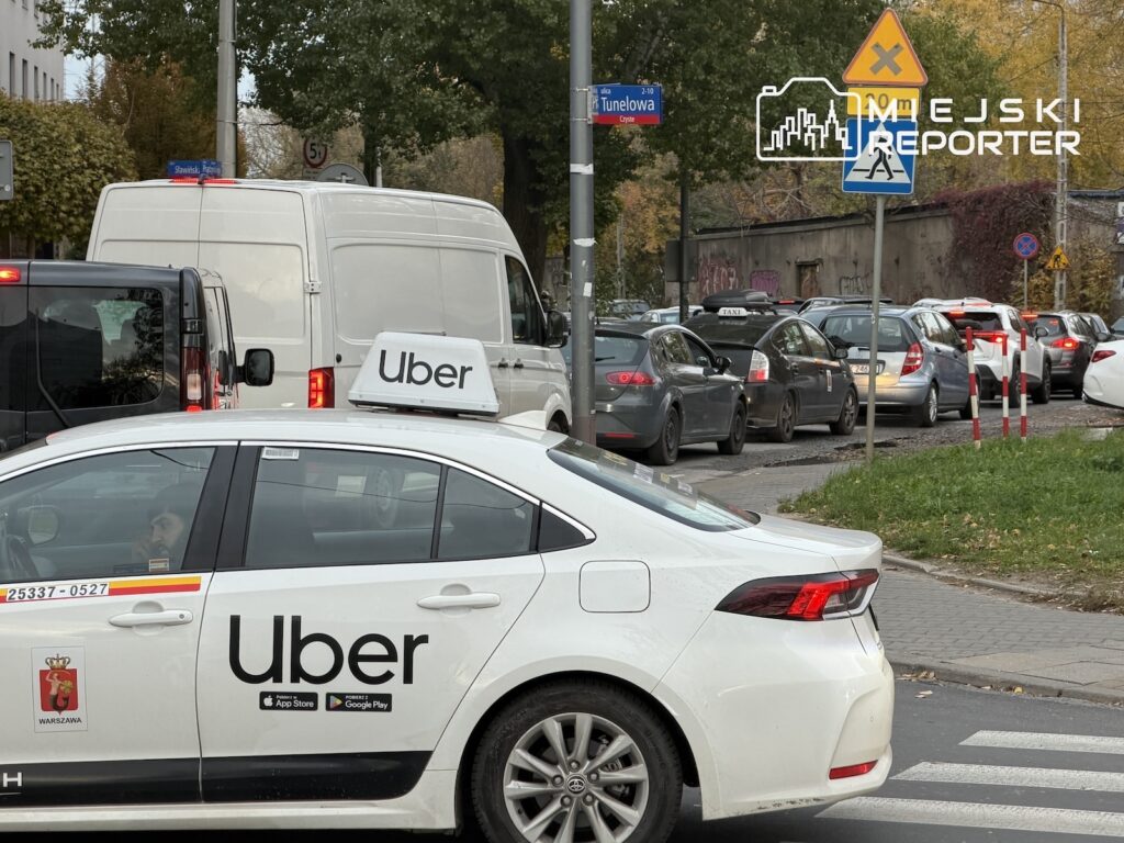 Biały samochód Ubera i sznur aut stoją w korku na ulicy przy przejściu dla pieszych.