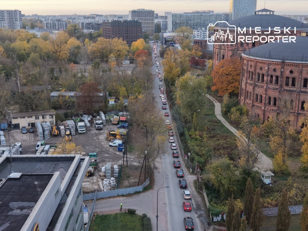 Długi sznur samochodów na wąskiej ulicy wśród jesiennych drzew, widziany z lotu ptaka.