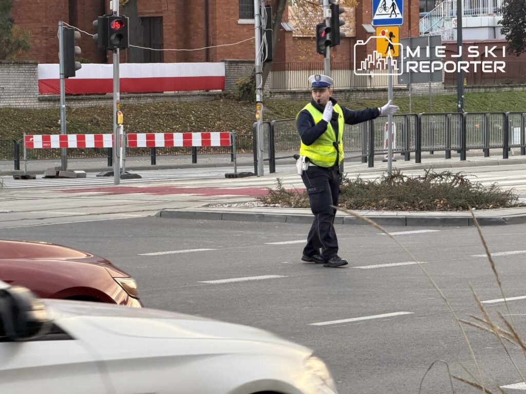 Policjant w kamizelce odblaskowej kieruje ruchem na skrzyżowaniu, przepuszczając samochody.